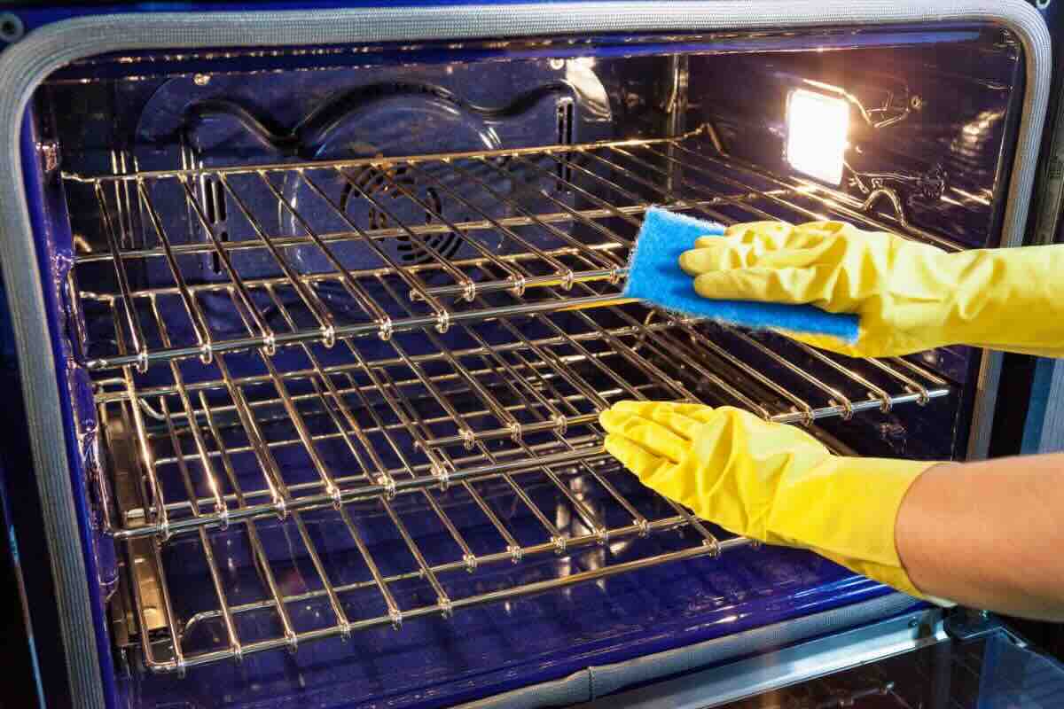 Mani con guanti gialli puliscono griglie forno con panno in microfibra blu