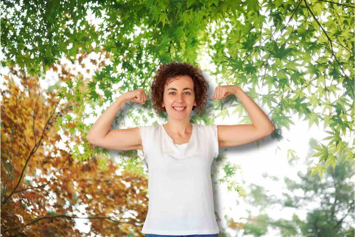 Donna braccia alzate, dietro di lei alberi foglie verdi e arancioni